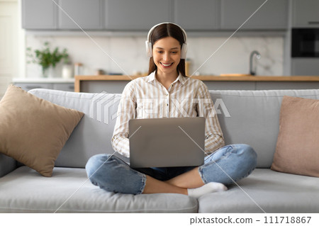 Smiling young woman with headphones using laptop on sofa 111718867