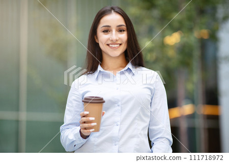 Happy arab young woman with smile, hold takeaway coffee cup, stands confidently outdoors 111718972