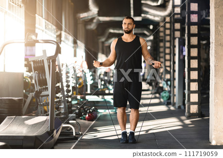 Cardio workout. Confident muscular man jumping with skipping rope in gym interior 111719059