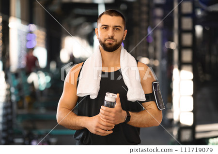 Muscular young man standing in gym with water bottle in hands Muscular young man standing in gym with water bottle in hands 111719079