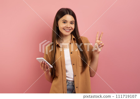 Cheerful teenage girl holding smartphone and making peace gesture Cheerful teenage girl holding smartphone and making peace gesture 111719107
