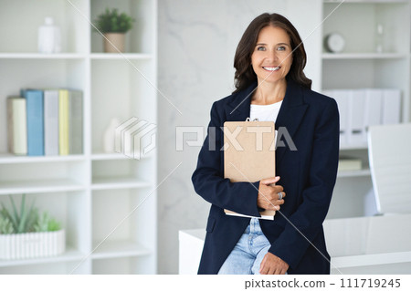 Confident mature woman posing in modern office, holding folder 111719245