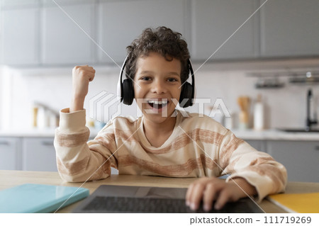 Cheerful black boy wearing wireless headphones using laptop computer and celebrating success 111719269