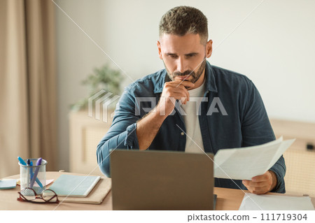 man looking at laptop with thoughtful face holding papers indoor man looking at laptop with thoughtful face holding papers indoor 111719304