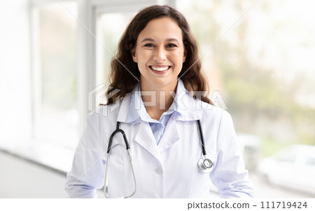 Cheerful millennial woman general physician standing by window 111719424