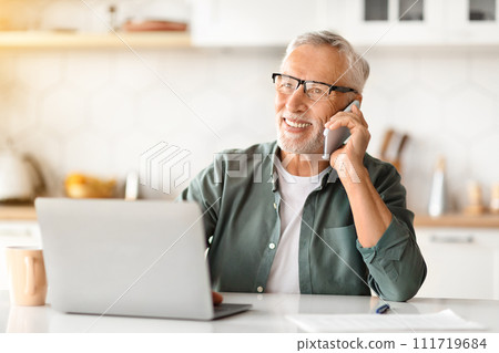 Smiling Elderly Gentleman Using Laptop And Talking On Cellphone In Kitchen 111719684