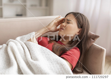 Uncomfortable young asian woman lying down on couch and touching temple 111720297