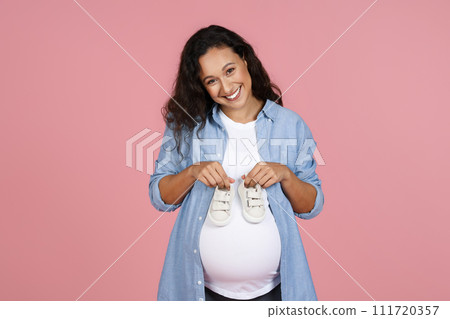 Joyful beautiful pregnant woman holding baby booties on her tummy 111720357