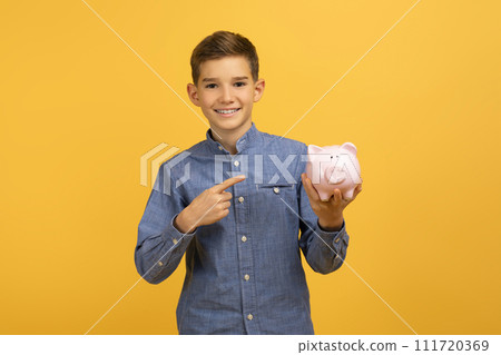 Happy excited teen boy pointing at piggy bank and looking at camera Happy excited teen boy pointing at piggy bank and looking at camera 111720369