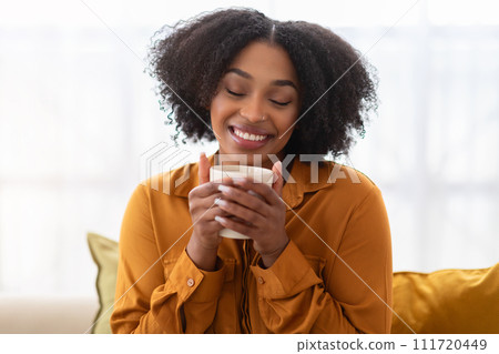 An enchanting young woman with a gentle smile and closed eyes inhales the aroma of her drink 111720449