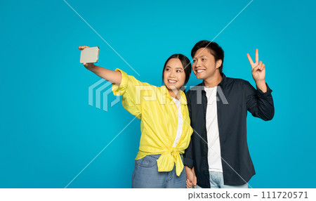 Cheerful woman in yellow taking a selfie with a smartphone while a man in dark attire poses with a peace sign 111720571