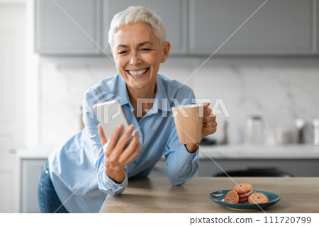 Happy senior woman using phone drinking her morning coffee indoors 111720799