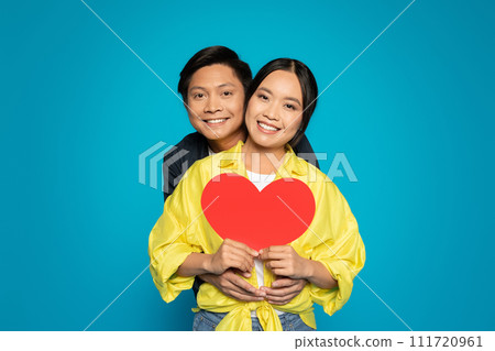 A radiant couple hugs joyfully, holding a large red heart together, with bright smiles and love 111720961