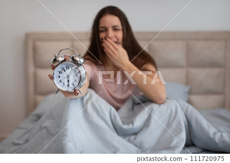 Sleepy woman in bed yawning, holding alarm clock in the morning 111720975