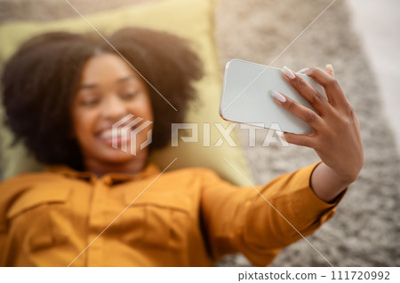 Smiling African American woman with curly hair takes a selfie while lying down 111720992