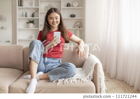 Relaxed young asian woman sitting on couch and looking at her smartphone 111721244