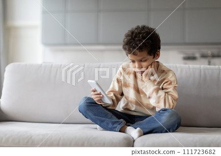 Little black boy sitting on sofa, looking disinterestedly at his smartphone 111723682