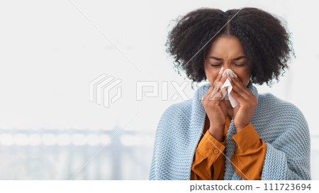 A young woman with curly hair appears unwell, blowing her nose with a tissue 111723694