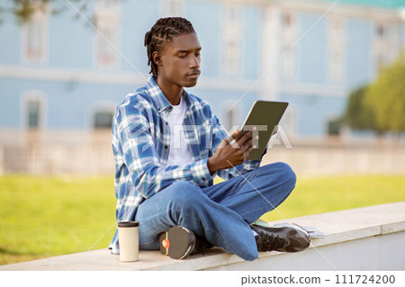 Young black man browses on tablet computer sitting on campus Young black man browses on tablet computer sitting on campus 111724200