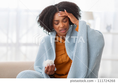 Distressed young woman with curly hair sitting on a couch, wrapped in a blue blanket Distressed young woman with curly hair sitting on a couch, wrapped in a blue blanket 111724268
