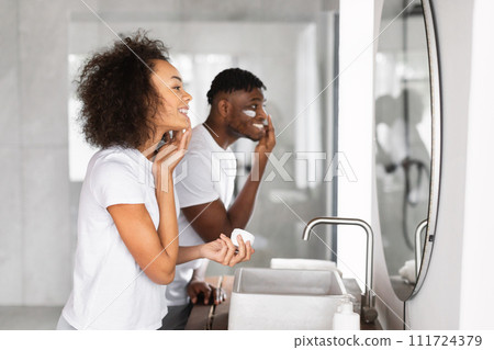 Loving black couple happily applying moisturizer near mirror in bathroom Loving black couple happily applying moisturizer near mirror in bathroom 111724379