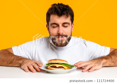 Young Caucasian man closes eyes smelling tempting burger, yellow backdrop 111724400