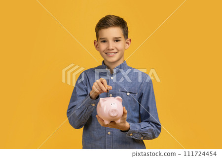 Smiling young boy in blue shirt depositing coins into pink piggy bank 111724454
