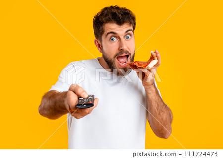 guy points remote controller savoring pizza slice on yellow backdrop guy points remote controller savoring pizza slice on yellow backdrop 111724473