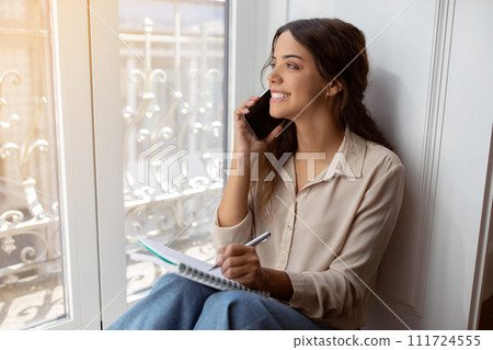Joyful young woman making phone call and taking notes by sunny window, 111724555
