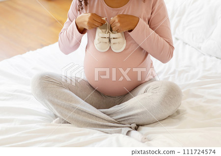 Pregnant lady sitting cross-legged on bed and holding pair of baby shoes 111724574