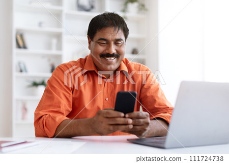Indian middle aged businessman texting on cellphone and using laptop Indian middle aged businessman texting on cellphone and using laptop 111724738