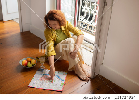 Thoughtful senior woman sitting on floor with glass of wine and map 111724924