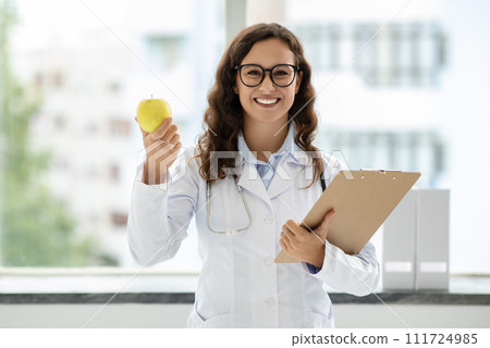 Friendly young woman doctor general physician showing apple Friendly young woman doctor general physician showing apple 111724985