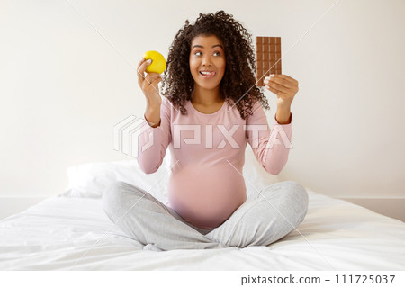 Cheerful black expectant mother sitting on bed, comparing apple to chocolate bar Cheerful black expectant mother sitting on bed, comparing apple to chocolate bar 111725037