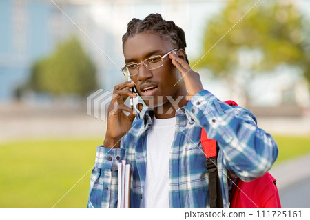 young black man holds backpack speaking by smartphone on campus 111725161