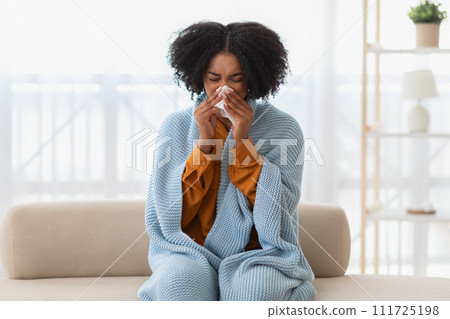 Young woman feeling sick, wrapped in a cozy blue blanket, sitting on the couch 111725198