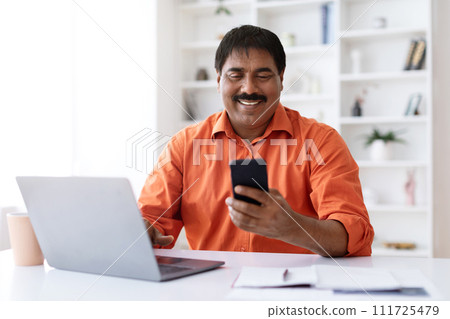 Cheerful mature indian man employee using laptop and smartphone Cheerful mature indian man employee using laptop and smartphone 111725479