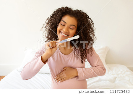 Beaming black expectant mother holding positive pregnancy test and embracing her belly Beaming black expectant mother holding positive pregnancy test and embracing her belly 111725483