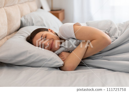 Content woman hugging blanket and smiling in bed 111725758