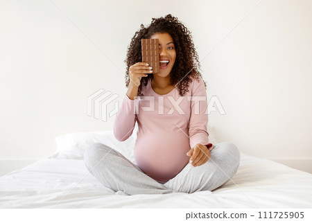 Laughing black pregnant woman covering one eye with chocolate bar Laughing black pregnant woman covering one eye with chocolate bar 111725905