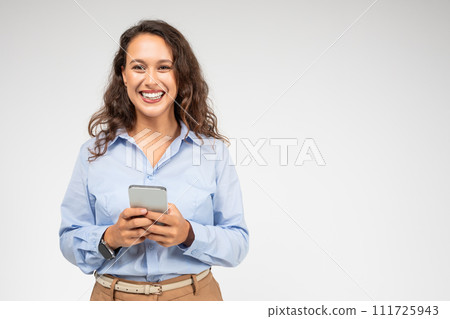 An engaging young woman with curly hair and a bright smile looks up from her smartphone An engaging young woman with curly hair and a bright smile looks up from her smartphone 111725943
