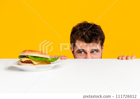 man peeking at tempting cheeseburger on plate against yellow background man peeking at tempting cheeseburger on plate against yellow background 111726012