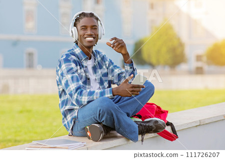 African student guy listening podcast with earphones and smartphone outdoors African student guy listening podcast with earphones and smartphone outdoors 111726207