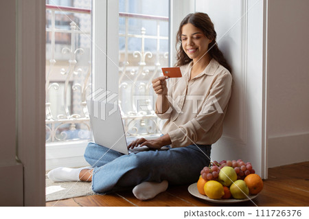 Smiling young woman sitting on floor with laptop and holding credit card Smiling young woman sitting on floor with laptop and holding credit card 111726376