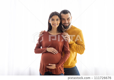 Portrait Of Pregnant Woman And Her Husband Embracing Near Window At Home Portrait Of Pregnant Woman And Her Husband Embracing Near Window At Home 111726676