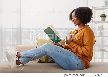 A relaxed woman with curly hair and a radiant smile sits cross-legged on a couch A relaxed woman with curly hair and a radiant smile sits cross-legged on a couch 111726750