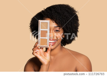 Happy young black woman holding concealer palette near face 111726783
