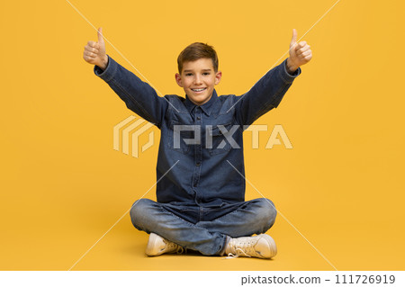 Cheerful Teen Boy Sitting On Floor In Studio And Showing Thumbs Up Cheerful Teen Boy Sitting On Floor In Studio And Showing Thumbs Up 111726919