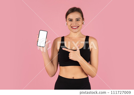 Smiling fitness woman in sportswear joyfully presenting blank smartphone screen 111727009