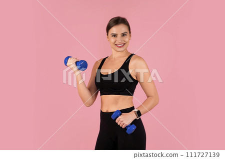 Energetic smiling woman exercising with blue dumbbells against pink studio background Energetic smiling woman exercising with blue dumbbells against pink studio background 111727139